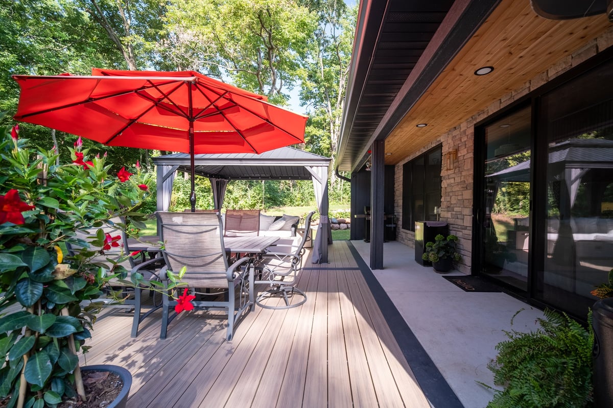 Modern patio with red umbrella, wooden deck, lounge chairs, and brick house with black doors surrounded by greenery