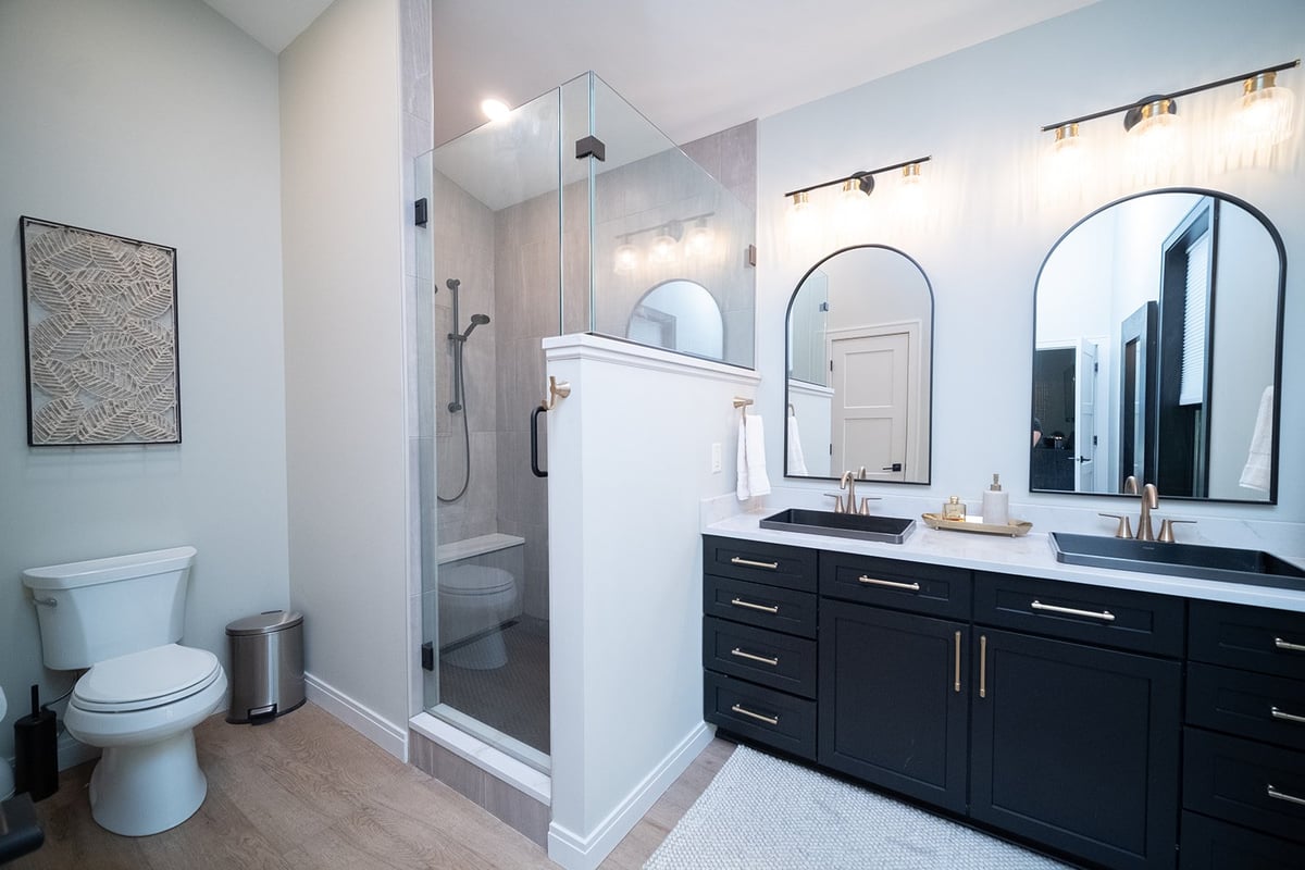 Modern bathroom with black double vanity, arched mirrors, glass shower enclosure, and toilet, featuring white walls and wood-look flooring.
